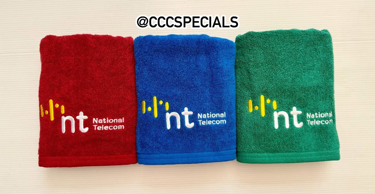 ผ้าขนหนูพรีเมียมสามสี ปักโลโก้ National Telecom ผ้าขนหนูสีแดง น้ำเงิน และเขียว ปักโลโก้ nt National Telecom วางเรียงกันบนพื้นผิวสีขาว
